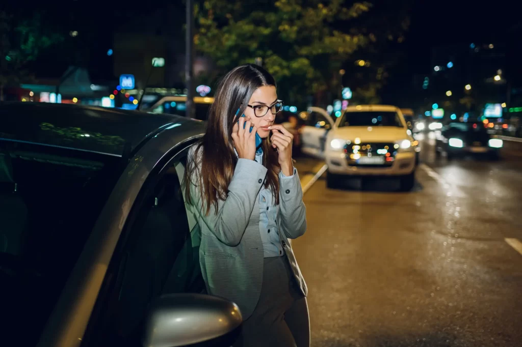 A woman standing beside a stranded vehicle on the roadside, using her phone to call a towing service for assistance.